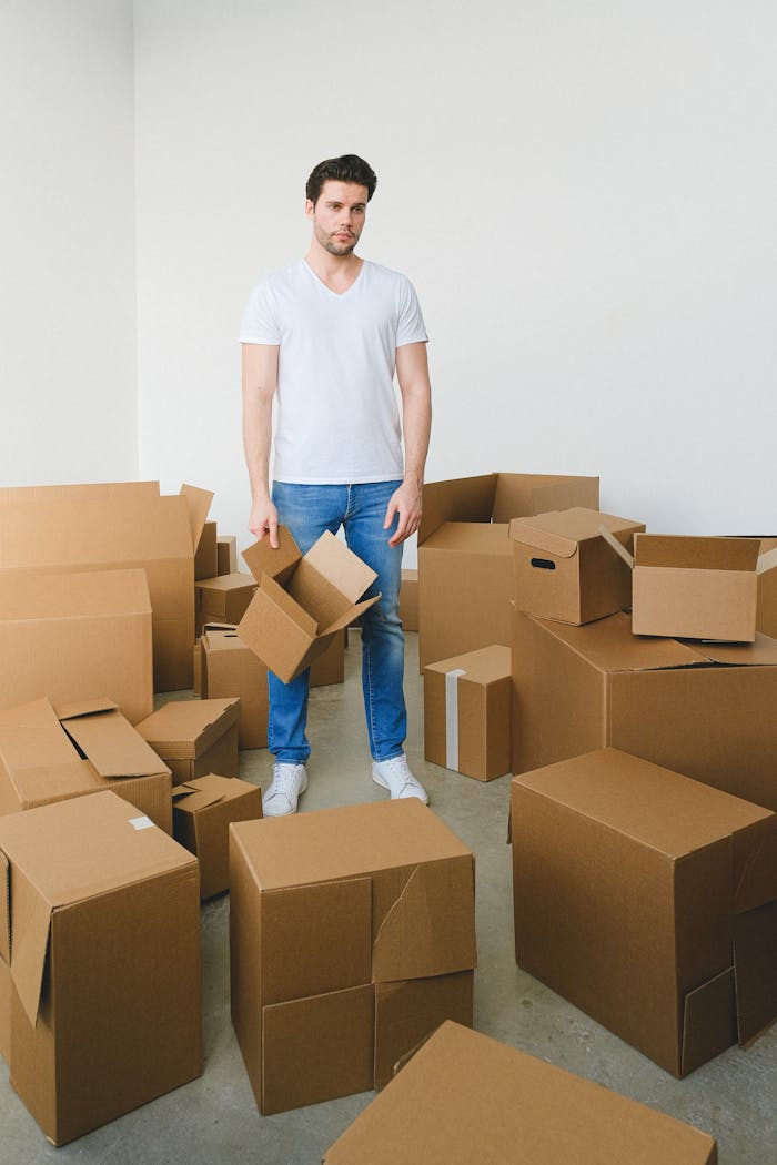 A thoughtful young man stands amidst cardboard boxes, symbolizing moving or relocation.