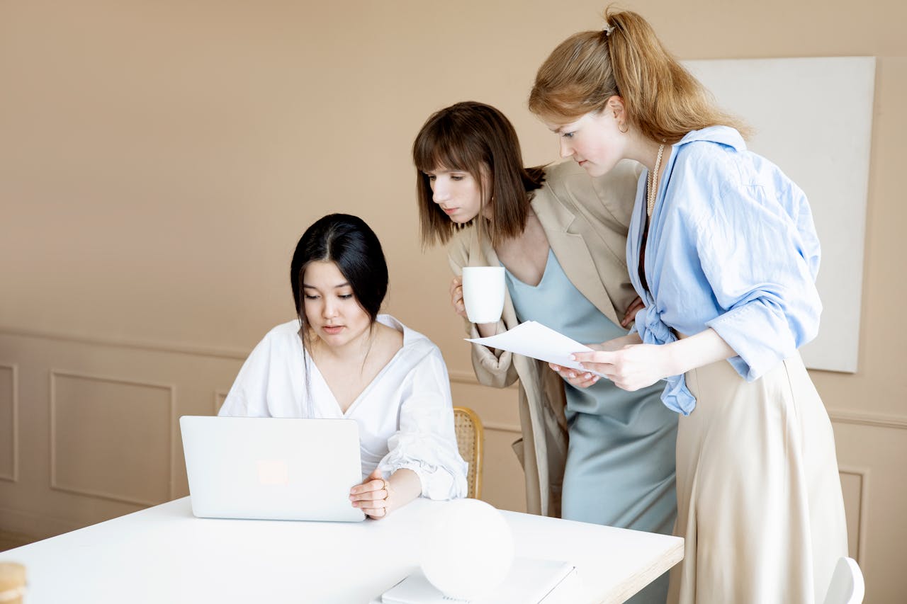 Three women collaborating in an office setting, using a laptop and discussing work.
